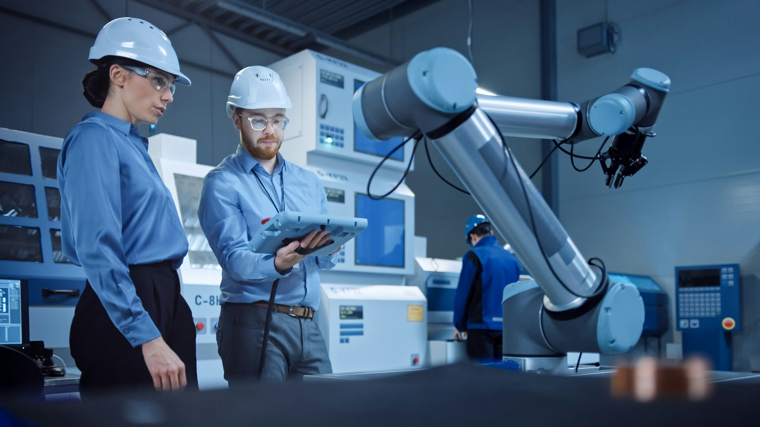 Two engineers in hard hats and safety glasses, one holding a tablet, observe a robotic arm in a modern industrial setting with machinery in the background.