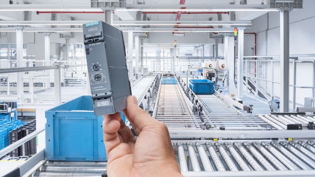Hand holding an industrial automation module in front of a modern conveyor system inside a manufacturing facility.
