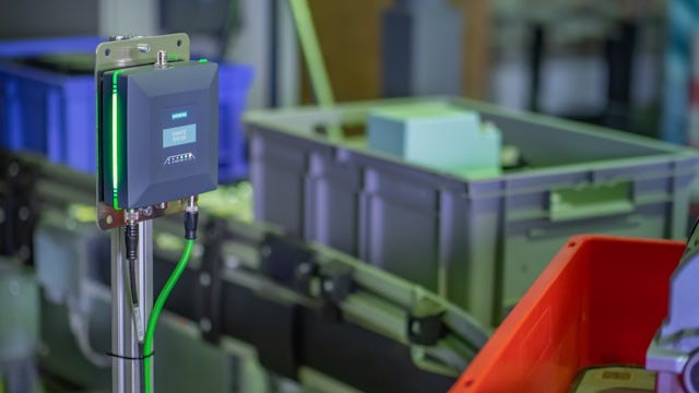 A Siemens industrial device with green indicator lights is mounted on a pole in a factory setting, with various bins and machinery blurred in the background.
