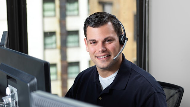 A smiling man wearing a headset with a microphone sits at a desk with multiple computer monitors, looking directly at the viewer with a city building visible through a window in the background.