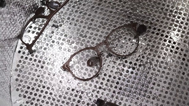 Eyeglass frames with water droplets rest on a perforated metal surface, suggesting a cleaning or manufacturing process.