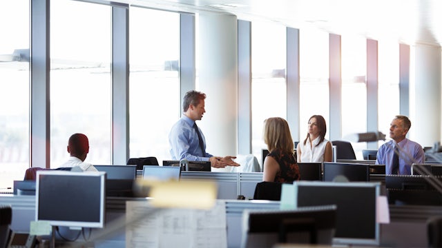 A group of colleagues in an office, sitting at desks while one colleague is standing and talking to them.