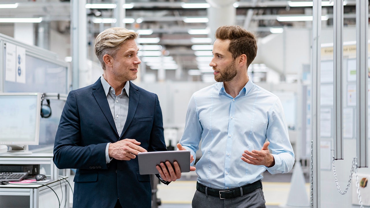 Two men walking in an office and discussing information on a tablet device.