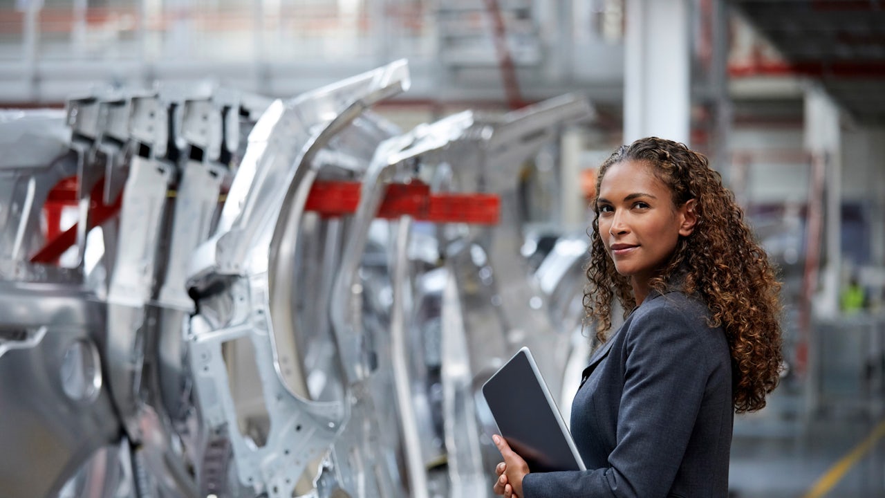 A woman wearing a business jacket and holding a tablet device stands in automotive factory line.