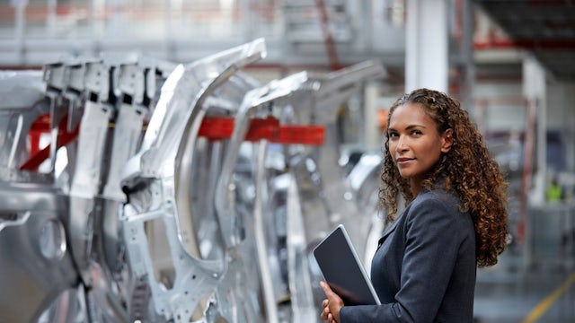 A woman wearing a business jacket and holding a tablet device stands in automotive factory line.