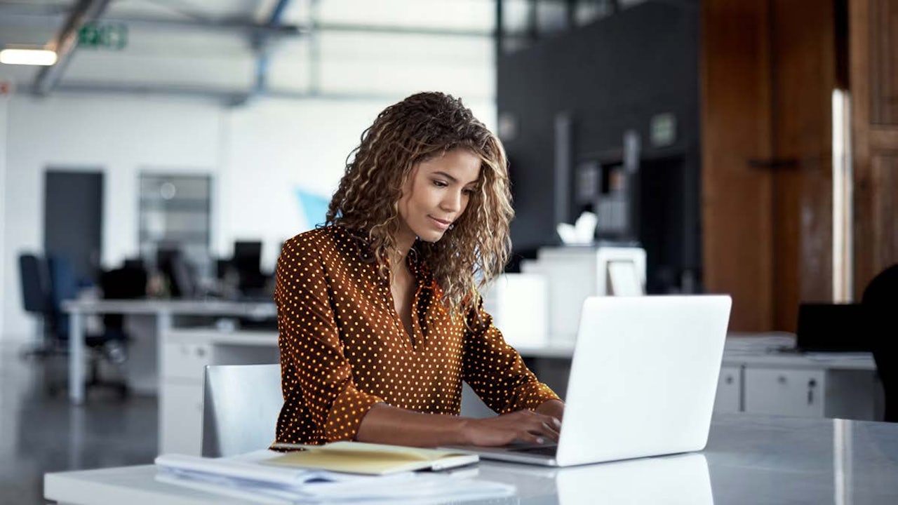 A woman in a brown blouse is working on a laptop in an office.