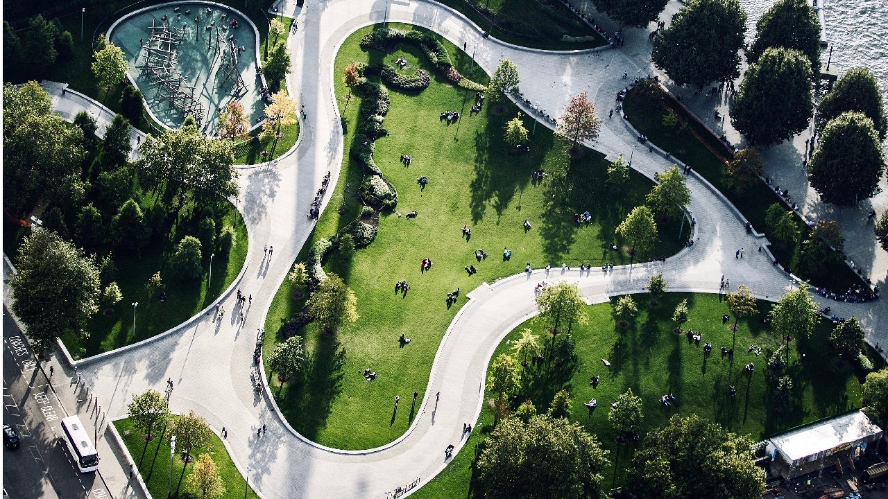An aerial view shows a winding path through a lush green park with trees and people, bordered by a body of water.