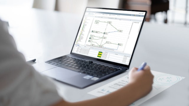 Close-up view of a laptop displaying an industrial network diagram as a person at a desk writes notes beside the device, illustrating digital engineering and network planning workflows.