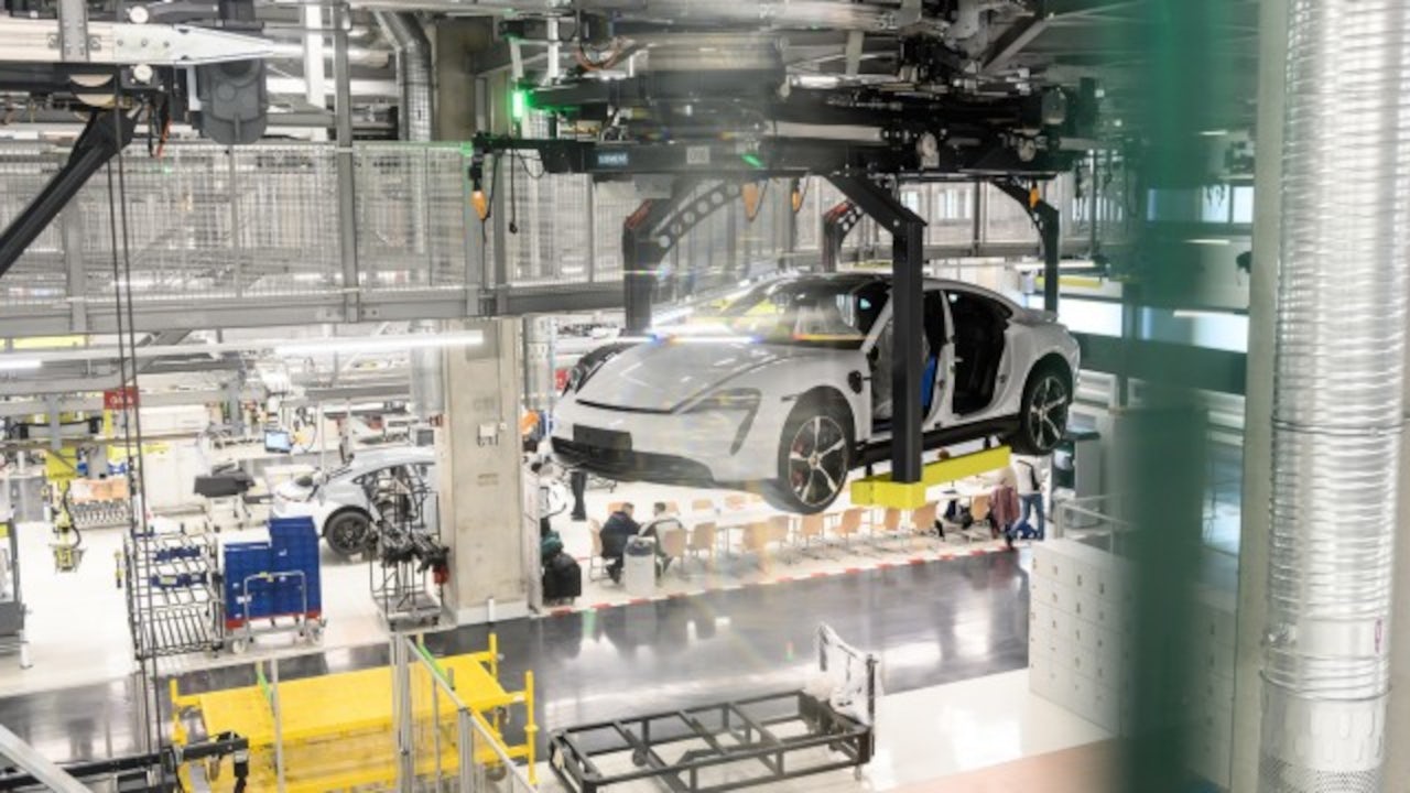 Automotive assembly line where a partially assembled white car is suspended by an overhead conveyor system inside a factory.