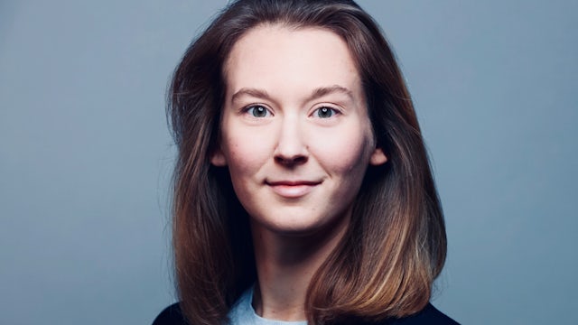 A woman with short hair and a serious expression is wearing a blue shirt and has a mole on her right cheek.