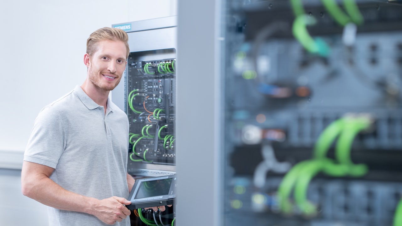 Industrial technician conducting network monitoring in front of a SCALANCE and RUGGEDCOM control cabinet, using a portable device to assess cabling and automation hardware inside a Siemens communication rack.