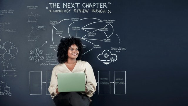 Young woman on a stool with an open laptop in front of a chalkboard.