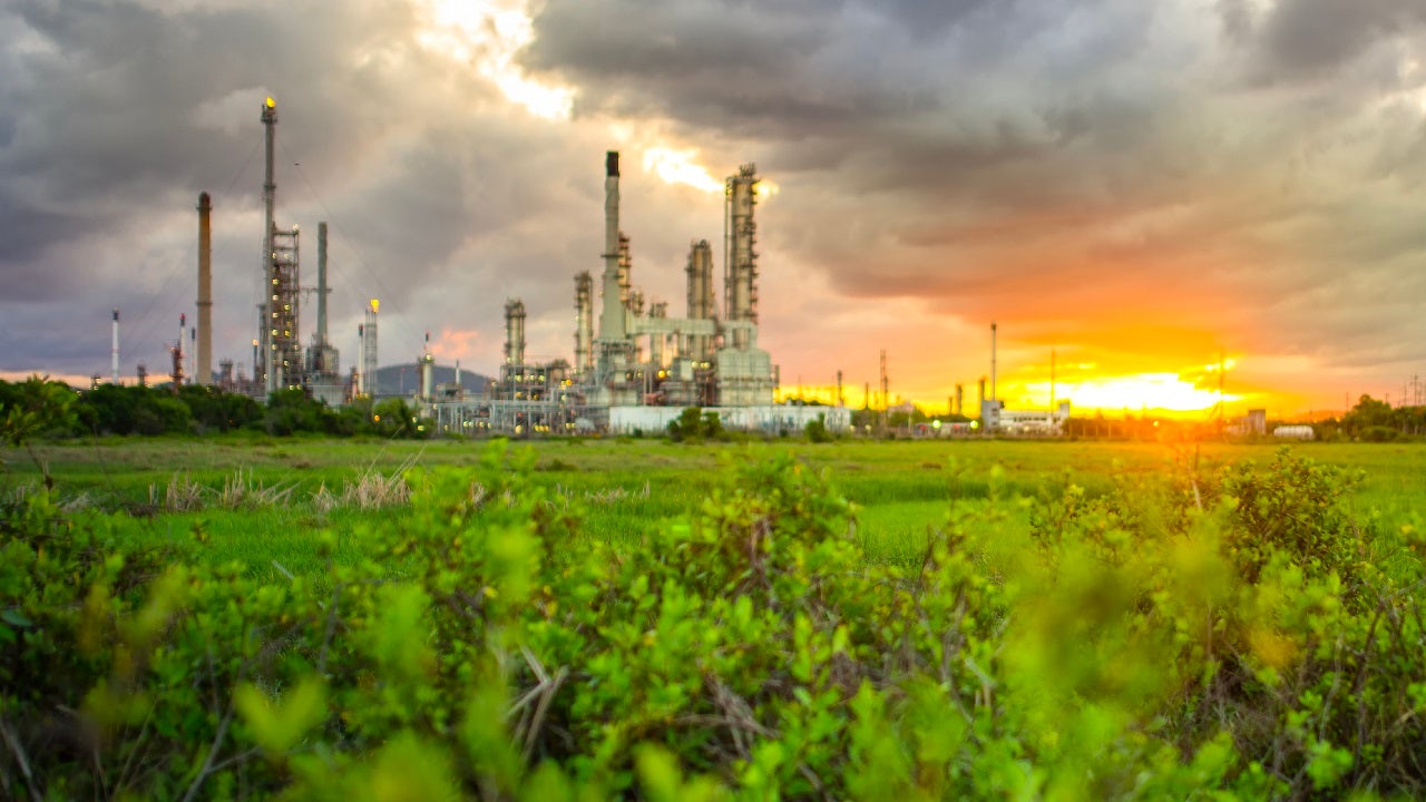 A large industrial plant with multiple smokestacks is silhouetted against a vibrant sunset over a field of green plants.