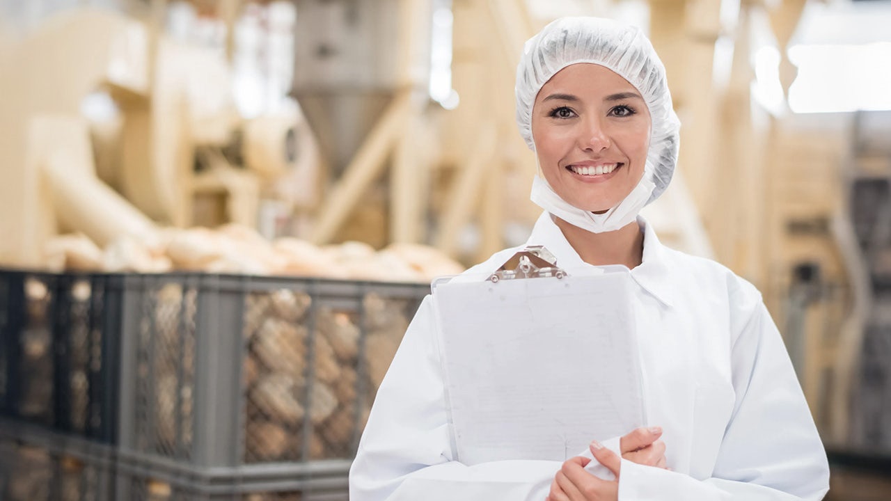 Smiling woman in white lab coat and hairnet holds a clipboard in a modern bakery representing food safety and production oversight.