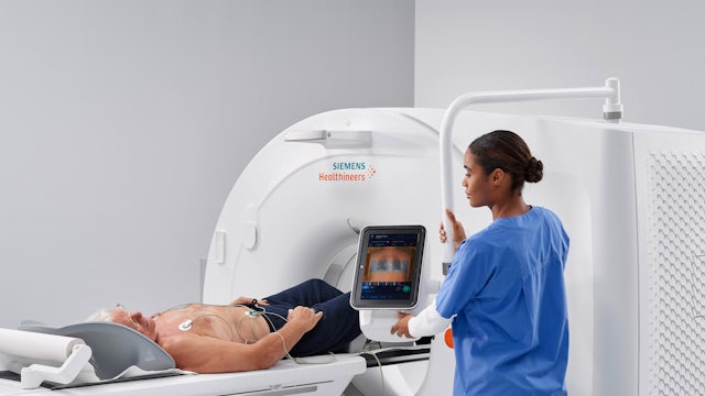 A medical professional in blue scrubs operates a Siemens Healthineers medical imaging machine with a patient lying on the bed, undergoing a scan.