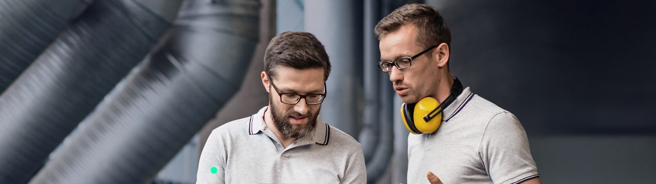 Two male engineers in an industrial setting, one holding a laptop, discussing data represented by green digital overlays.