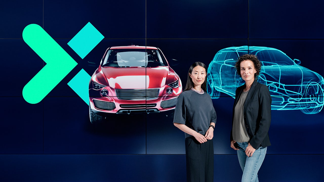 Two women standing in front of a dark blue background with a visualization of a car and its digital twin alongside the Xcelerator logo.