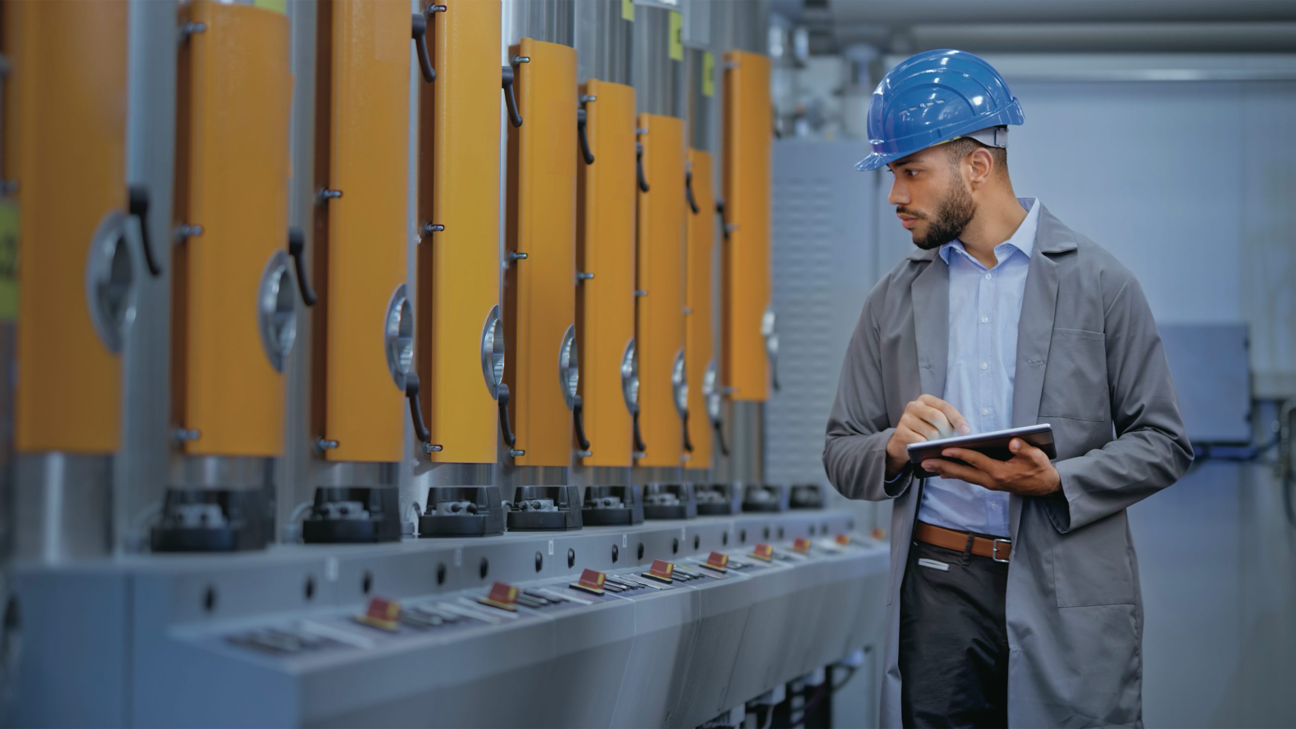Man wearing a hard hat and holding a tablet inspecting manufacturing machines in an industrial setting.