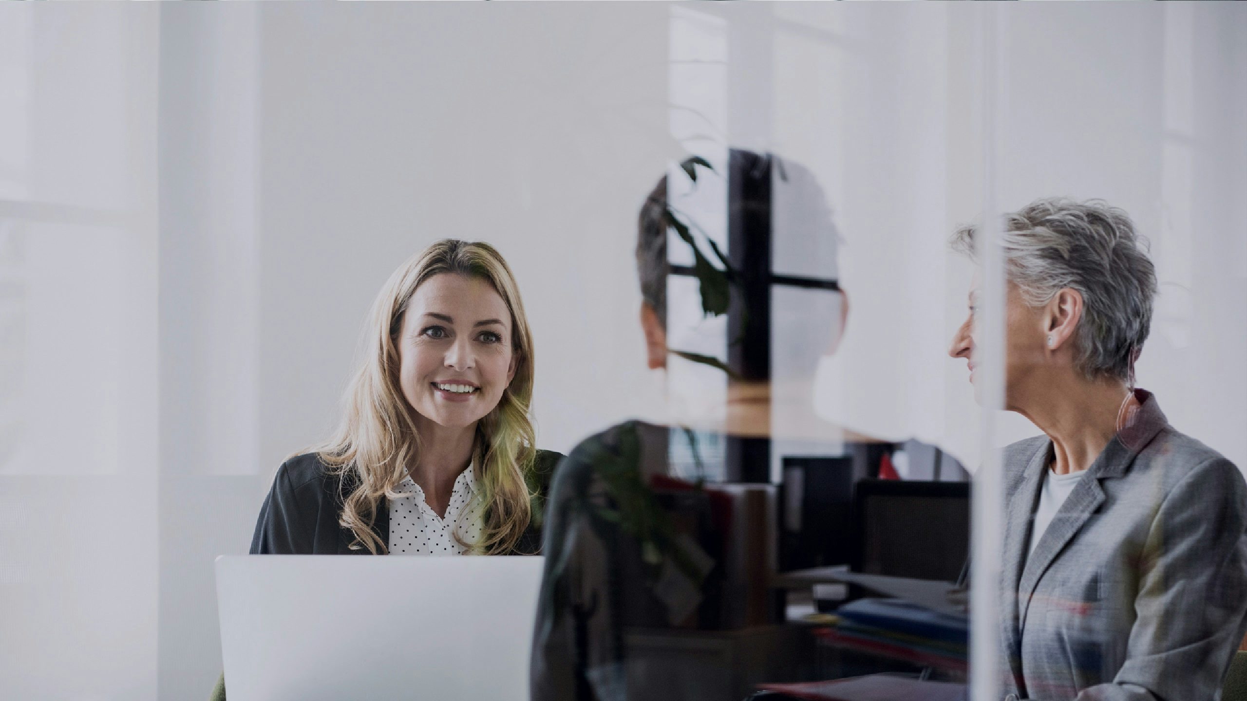 Three business professionals, two women and one person with their back to the viewer, are seated around a white circular table in a modern office, with a smiling blonde woman on the left and reflections from a glass partition visible.