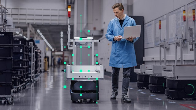 A man in a lab coat uses a laptop next to an automated guided vehicle with green data overlays in a factory setting.