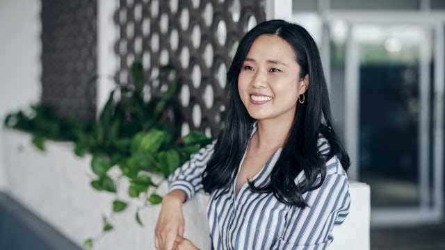 A smiling, professional woman leaning against a wall in a building with greenery behind her.