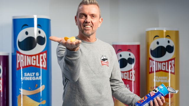 A man in a gray Pringles sweatshirt smiles at the camera, holding a small Pringles can in one hand and a few Pringles chips in the other, with giant Pringles cans in various flavors behind him.