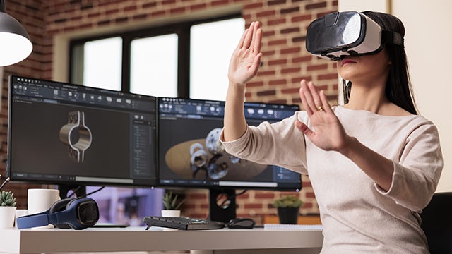 A woman wears a VR headset in front of a computer monitor displaying a machine part