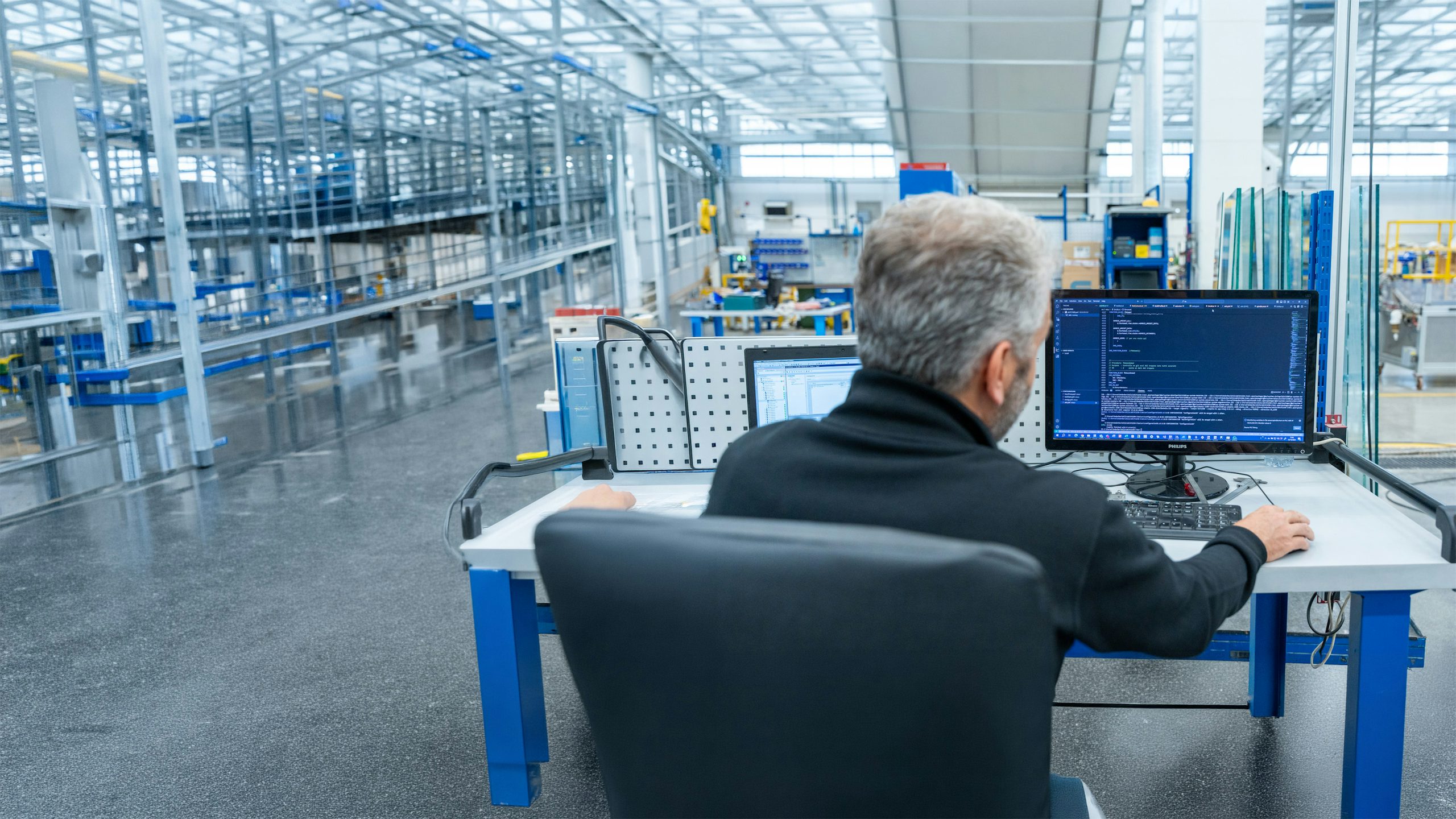 Over-the-shoulder view of a grey-haired engineer sitting at a desk in an industrial facility. He is looking at a Philips monitor displaying lines of Structured Control Language (SCL) code within a modern development environment, with a factory floor visible in the blurred background.