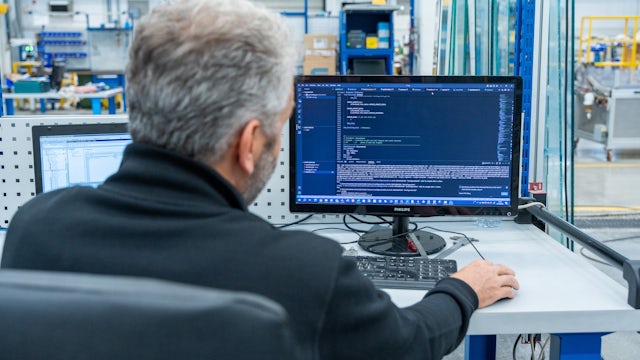 Over-the-shoulder view of a grey-haired engineer sitting at a desk in an industrial facility. He is looking at a Philips monitor displaying lines of Structured Control Language (SCL) code within a modern development environment, with a factory floor visible in the blurred background.