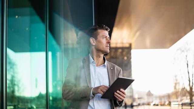 A businessman stands under a building awning with a tablet.