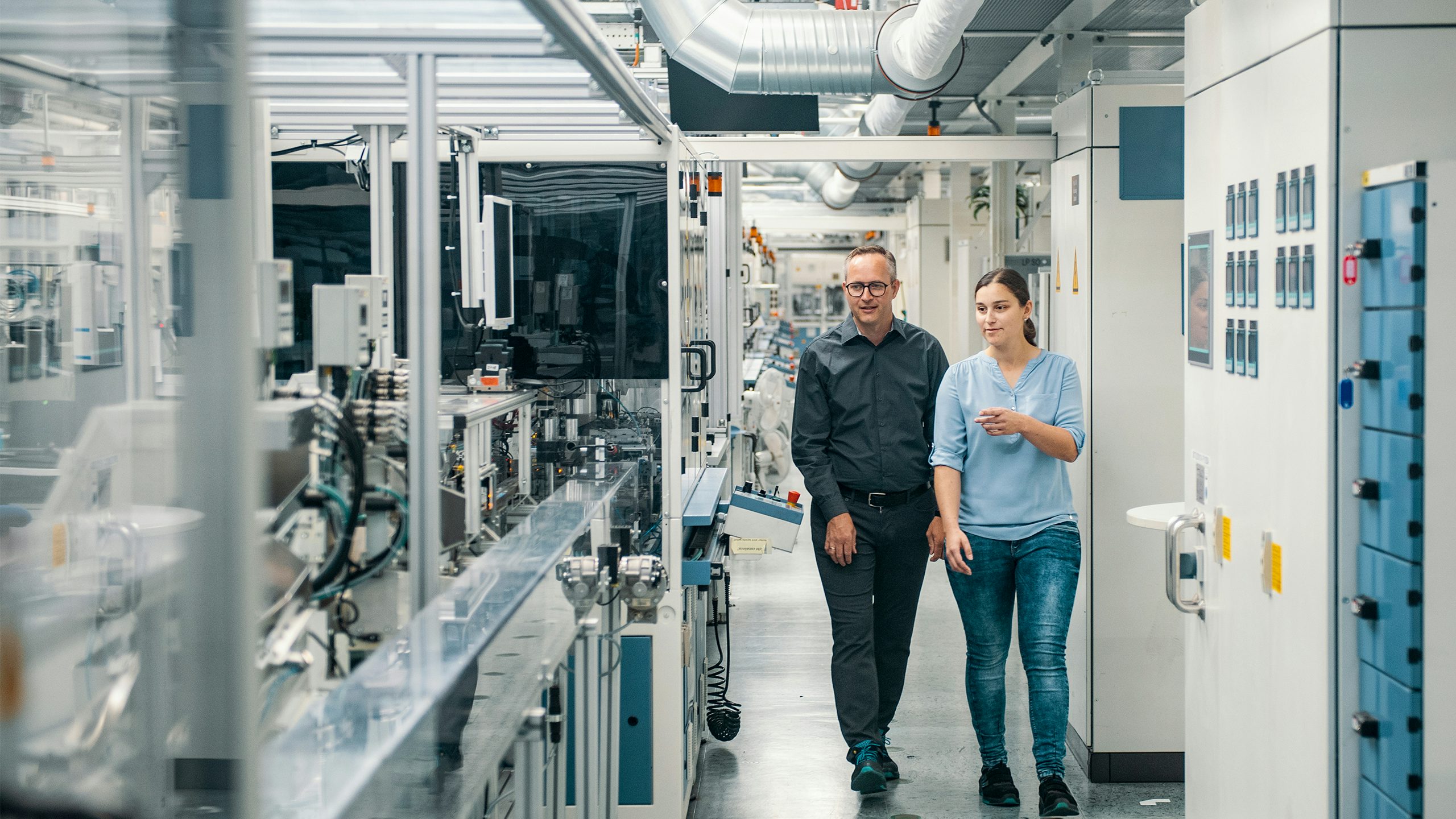 A man and a woman walk through a modern industrial facility, passing by production machinery on the left and large control panels on the right. The woman is gesturing as they talk.