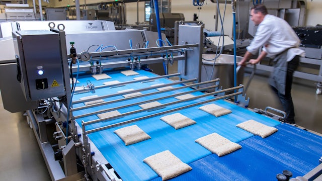 Man pushes cart next to industrial bakery machine, with raw products arranged in rows on blue conveyer belt. 