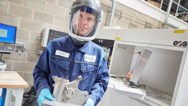 A man wearing a uniform and a helmet, while holding a 3D printed object in a manufacturing setting.