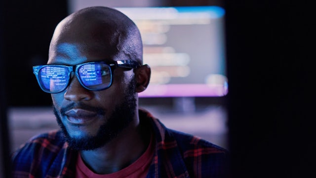 A focused male IT professional, wearing glasses that reflect lines of code, works intently in a dimly lit server room or data center control environment.