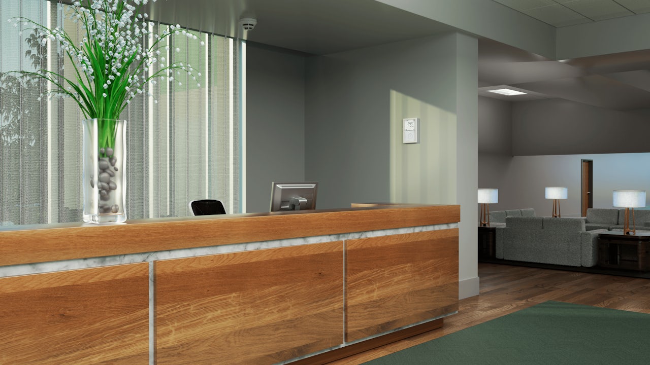 A modern reception desk with a large floral arrangement, computer monitor, a thermostat on the wall, and a waiting area with sofas and lamps.