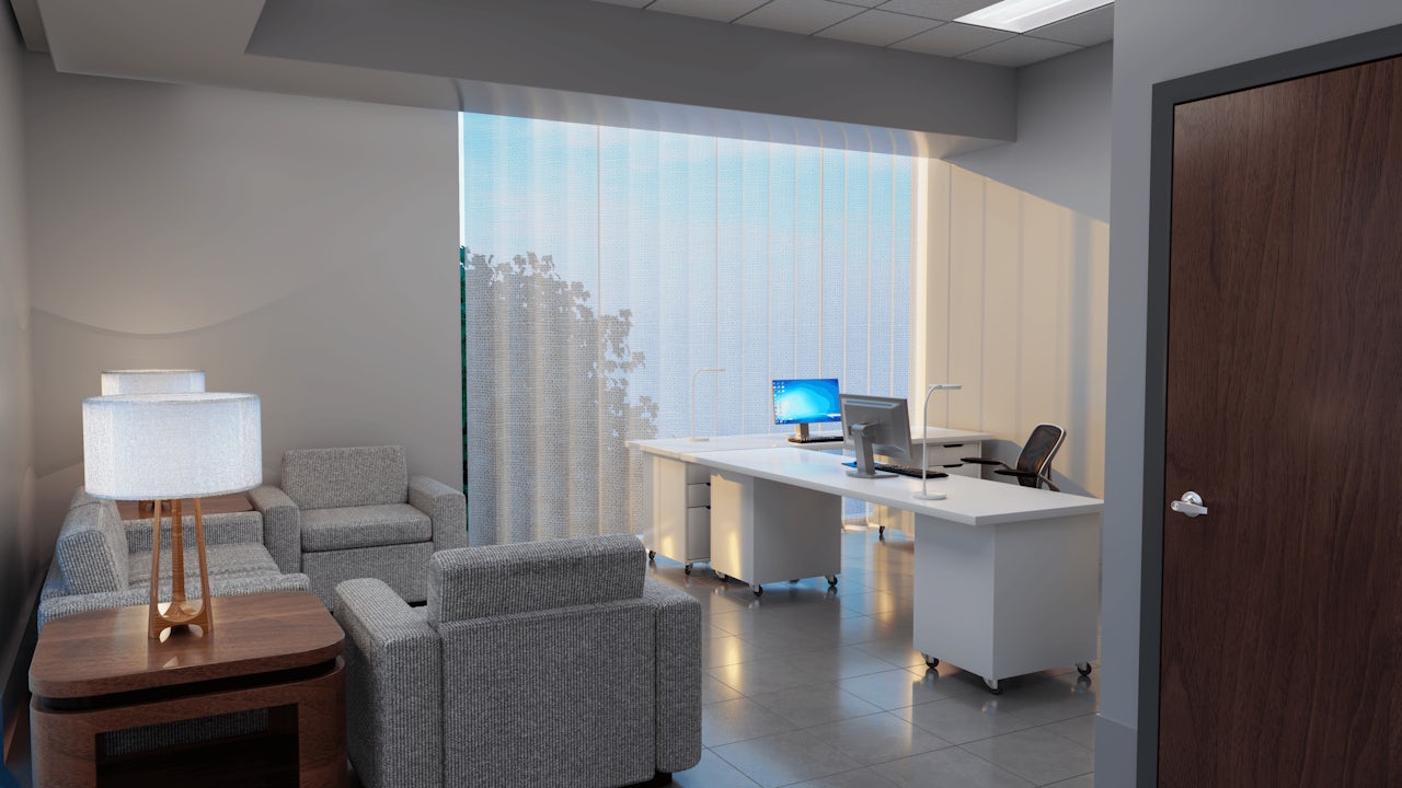 Office with a waiting area featuring gray armchairs and a wooden side table with a lamp, and a workspace with white desks and computers by a large window.
