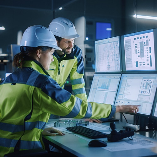 Two engineers in a control room wearing safety gear reviewing data and diagrams on multiple monitors.