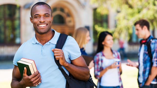 A university student, wearing a backpack, holding a book and smiling, while his classmates are in the background talking.