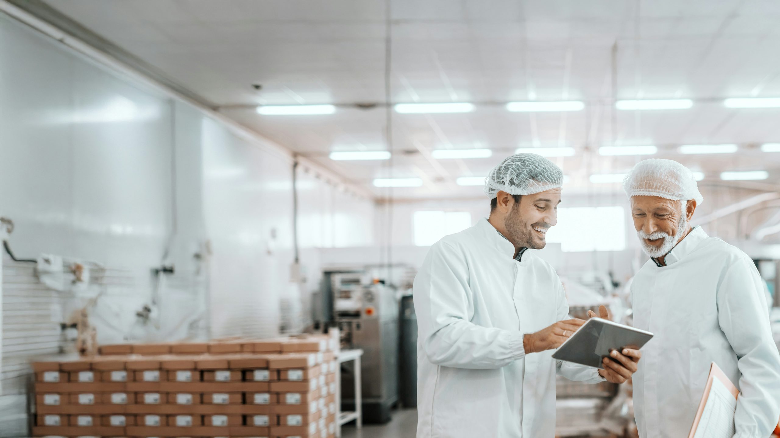Two manufacturing professionals wearing protective lab coats and hair nets review information on a tablet together inside a production facility.