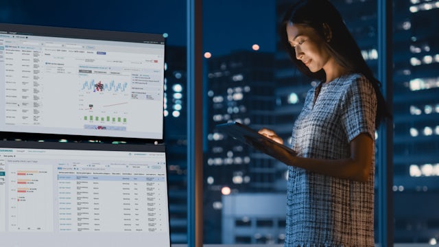 Woman uses tablet next to two large glowing monitors.