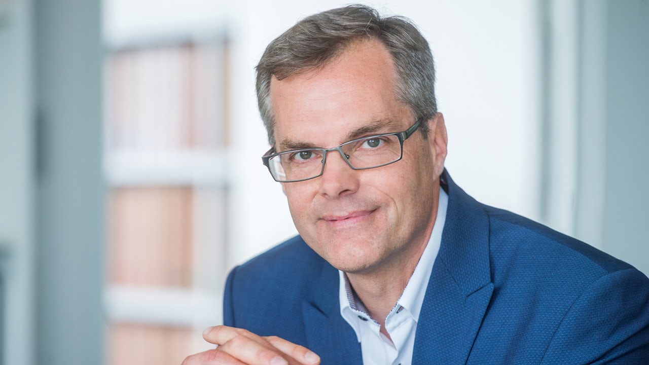 A man in a suit is standing in front of a Siemens building, looking at the camera.