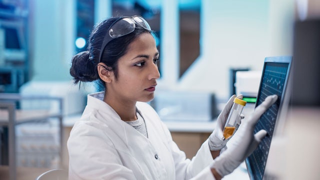 A female scientist is standing in a lab, wearing a lab coat and safety goggles, with a digital tablet in her hand. 