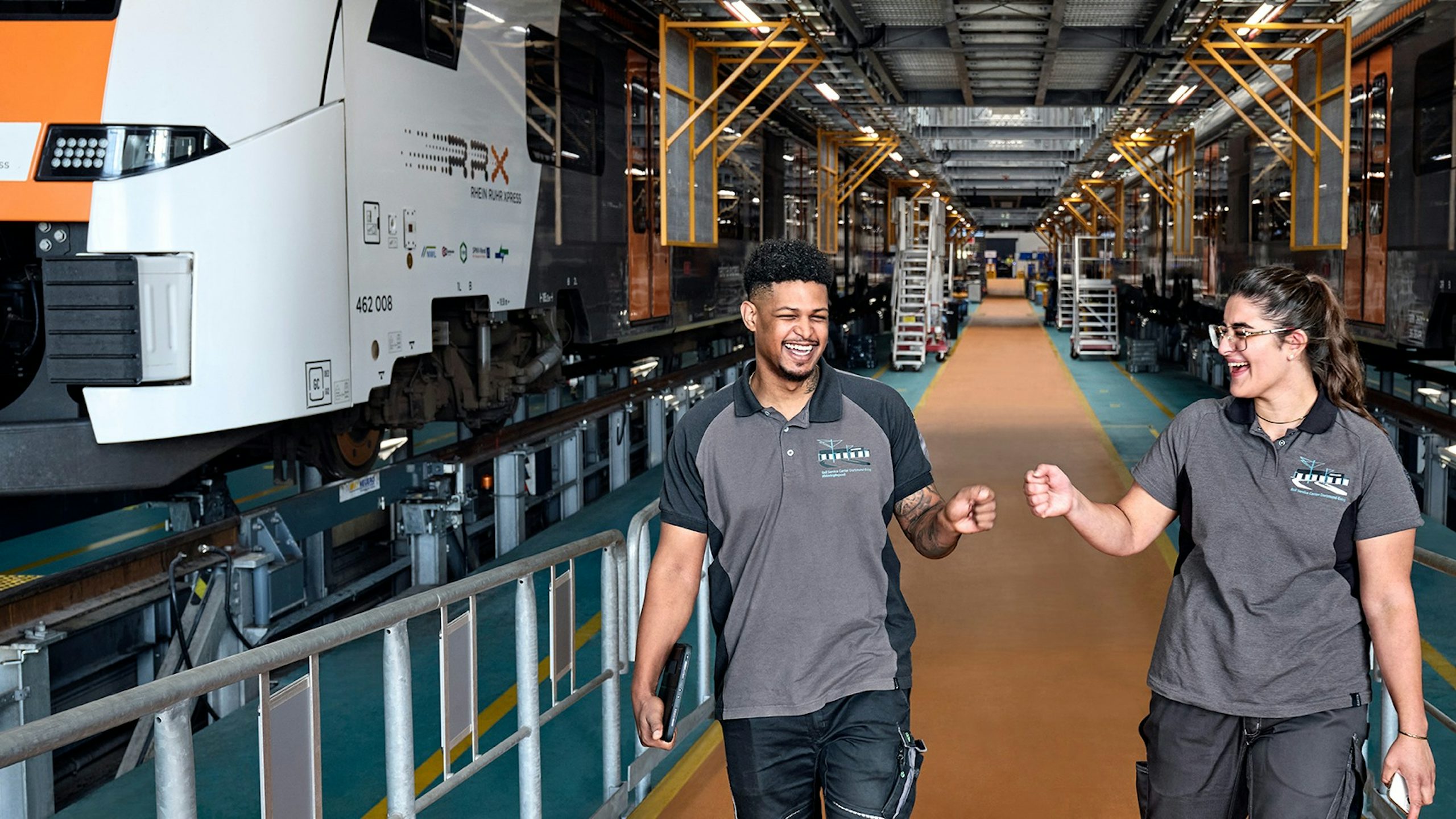 Two rail technicians walking and talking inside a train maintenance facility with trains on either side.
