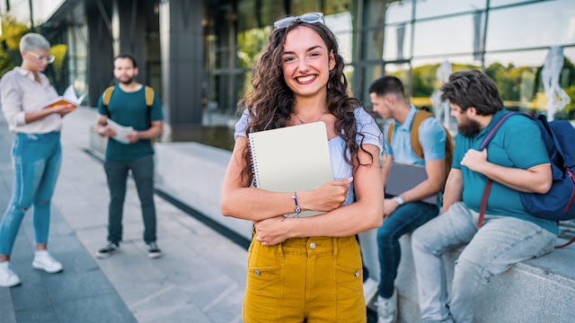 Female student smiling while holding books and a laptop on a university campus