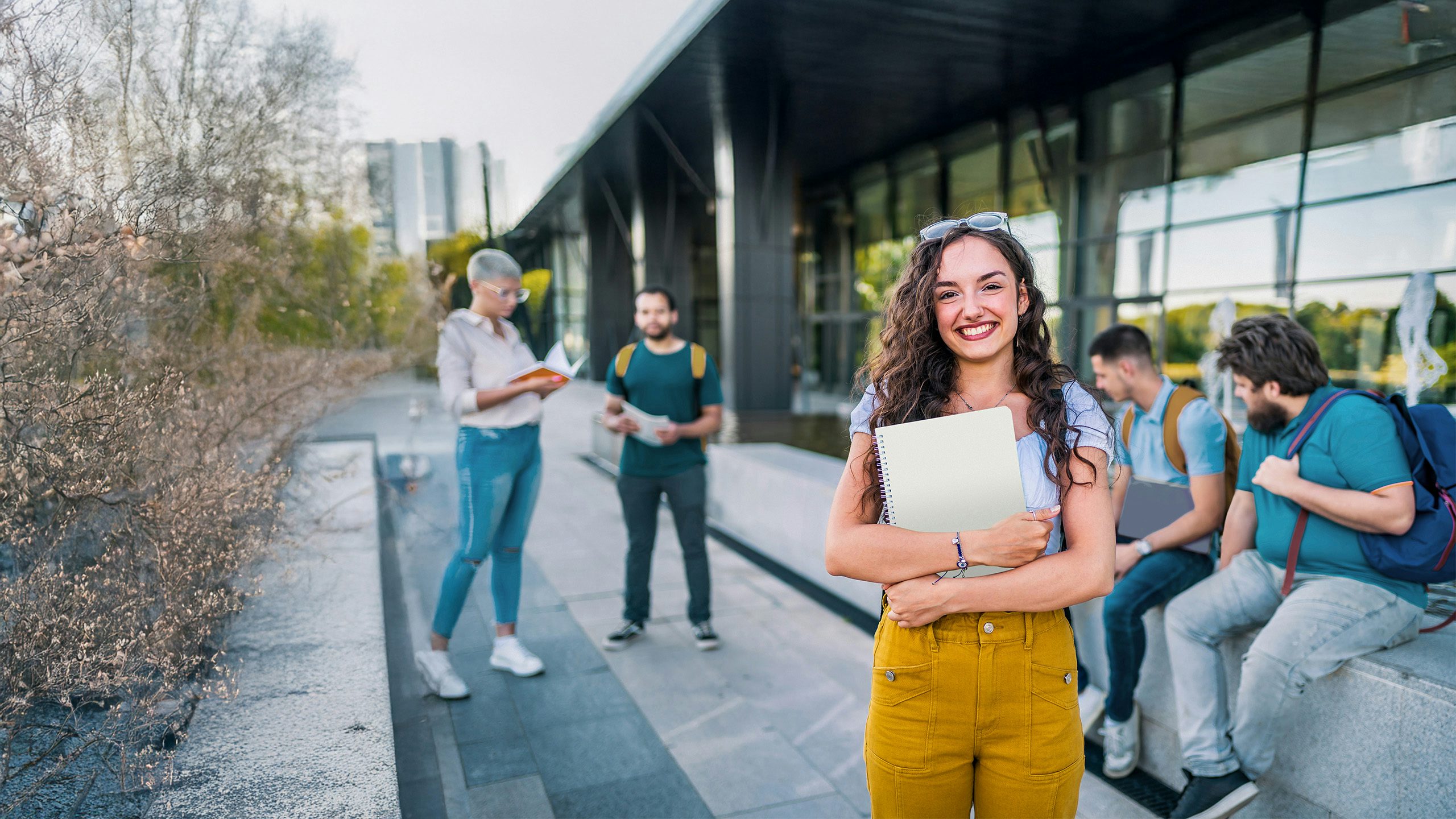 Young woman with curly hair smiling while holding books and a laptop on a university campus