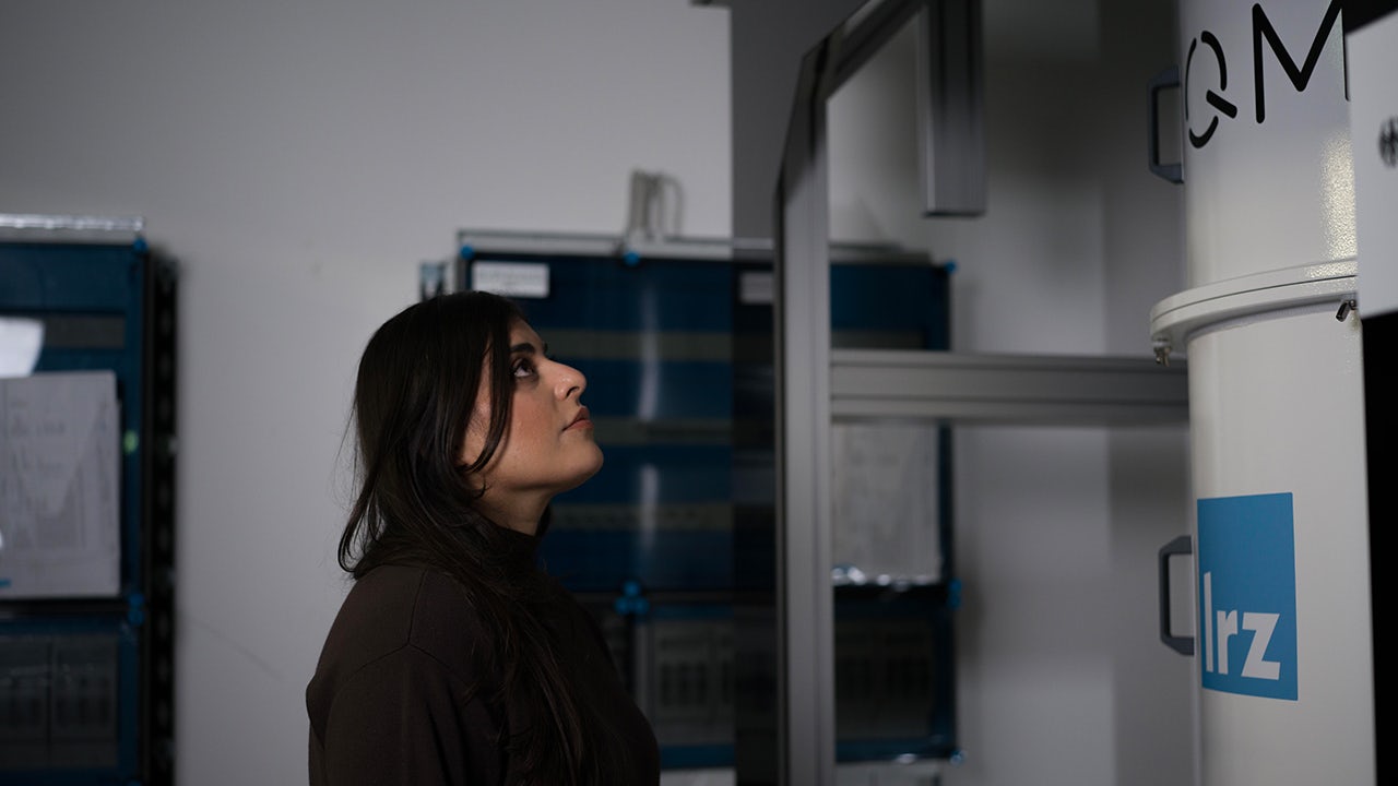 A woman in a factory floor discussing quantum computing with a large screen behind her.