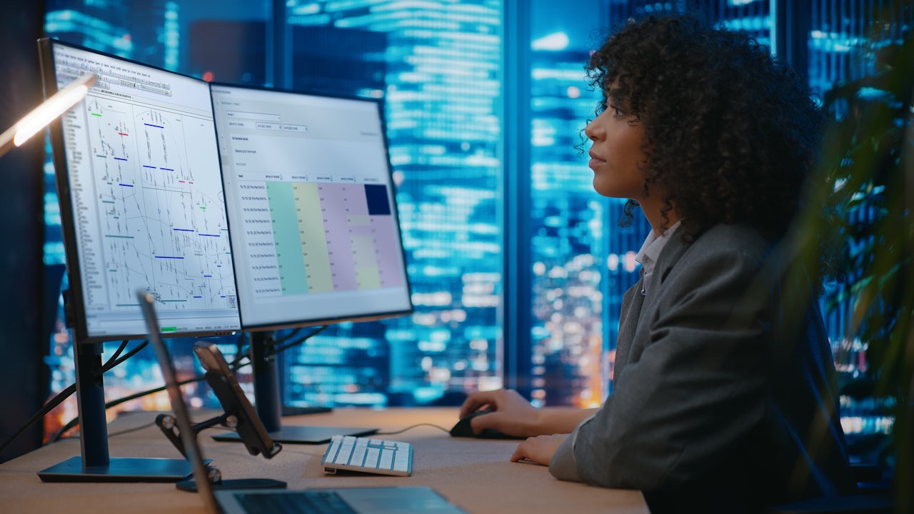A woman looks at a program on a computer screen at night, with skyscrapers behind her.  