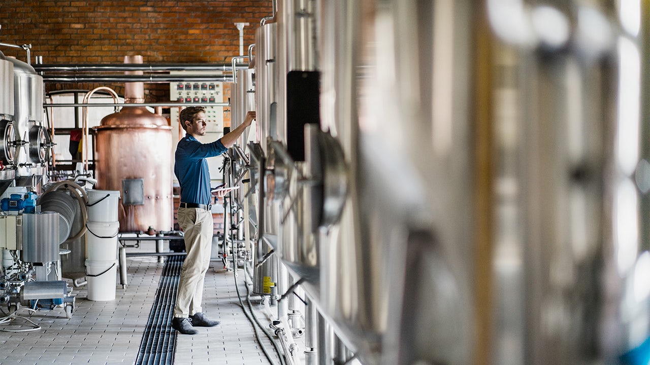 A man is working in a brewery, standing in front of a large metal tank, wearing a white shirt and a hat.