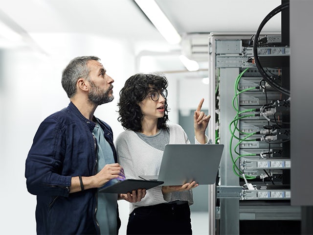 Two IT professionals, a man with a tablet and a woman with a laptop, inspect a server rack in a data center.