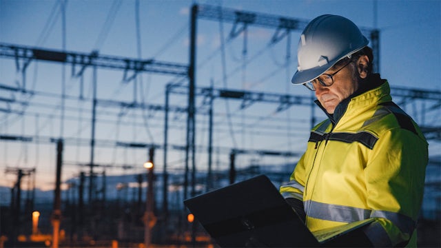 A worker in safety gear stands before a power plant, wearing a hard hat and a safety vest